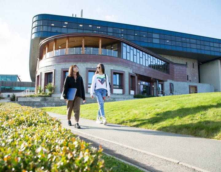 Two students walking past a large building on Penryn Campus