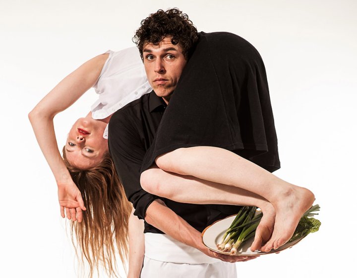 A male dancer stood upright with the female dancer on his shoulder and round his neck holding a silver tray of spring onions