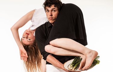 A male dancer stood upright with the female dancer on his shoulder and round his neck holding a silver tray of spring onions