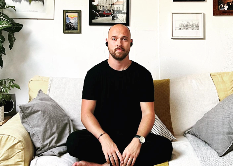 Graduate Thomas Young sitting cross-legged on a sofa with a wall of artwork and photos behind him