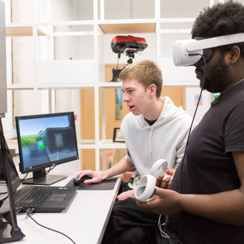 A student wears a VR headset while the other looks at a computer screen